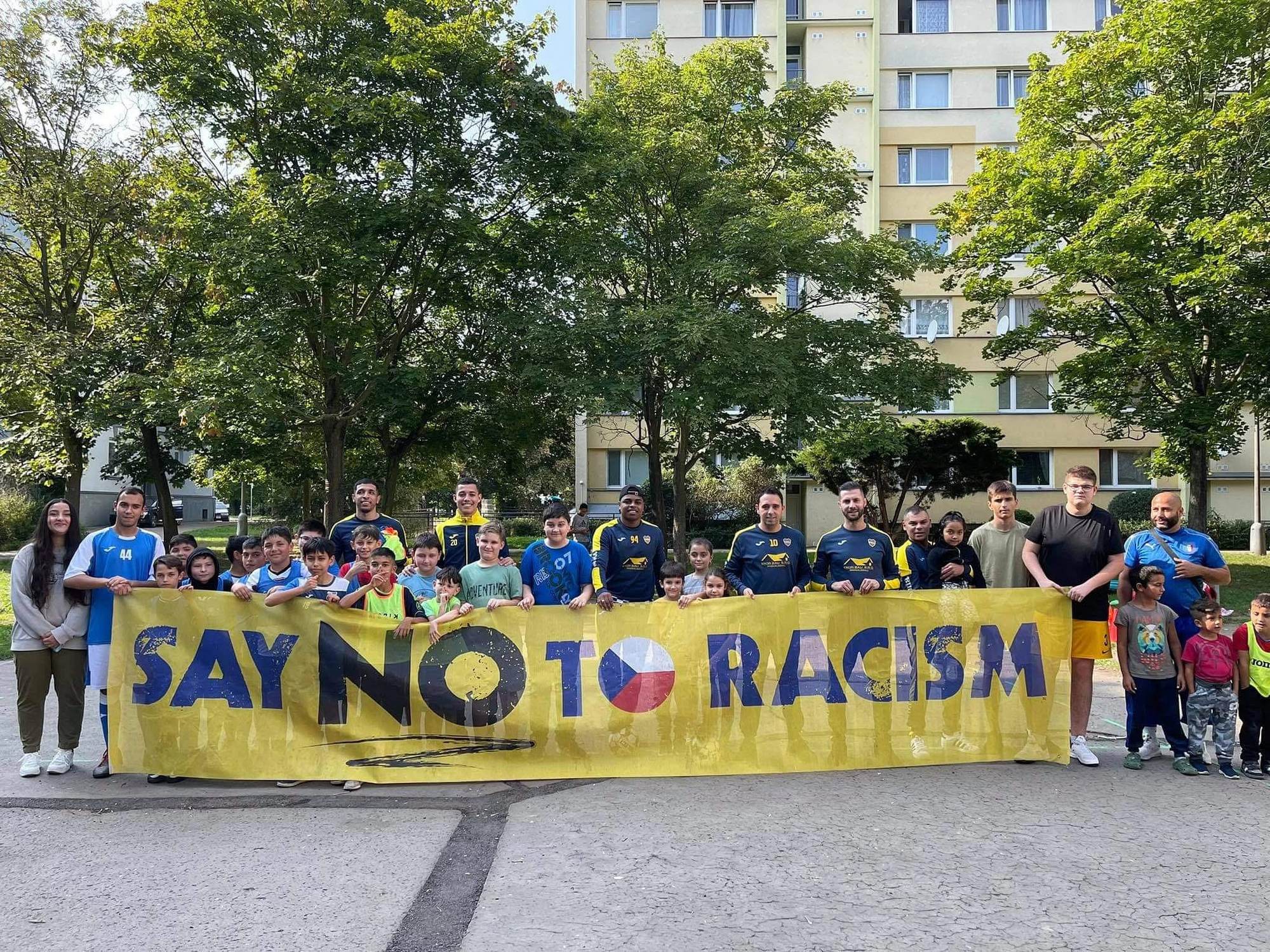 Děti a trenéři s transparentem Say no to racism před panelákem