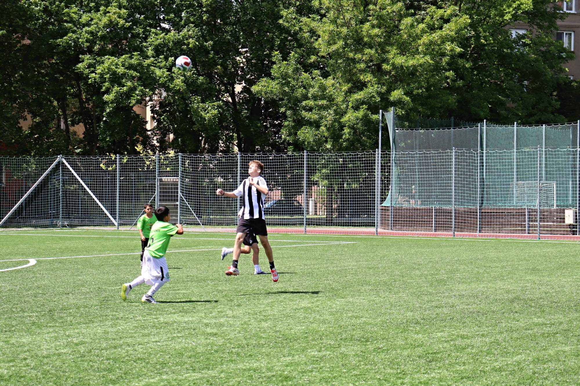 Kluci v zeleném a pruhovaném dresu hrají o vysoký míč na hřišti s umělou trávou